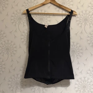 Svart korsett med dragkedja - Snygg svart korsett med dragkedja framtill och justerbara axelband. Perfekt för att skapa en stilren och elegant look. Passar bra till både vardag och speciella tillfällen.