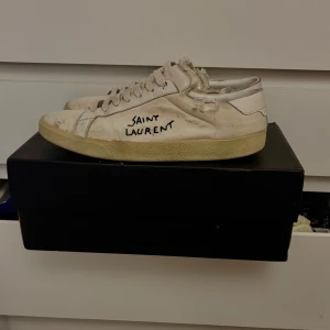 Vita Saint Laurent skor - Säljer ett par vita sneakers från Saint Laurent. Skorna har en klassisk design med snörning och en något sliten look. Perfekta för en avslappnad stil. Det medföljer kvitto, dustbag och box. Prisförslag✅
