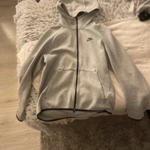 Grå huvtröja från Nike - Säljer en grå huvtröja från Nike med dragkedja och huva. Tröjan har långa ärmar och en liten Nike-logga på bröstet. Perfekt för en avslappnad stil. En gammal verision av Nike tech :)