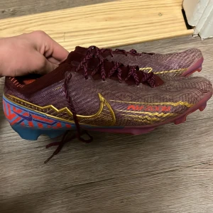 Nike mercurial Mbappe 2020 elite  - Säljer ett par unika fotbollsskor från Nike med en mix av lila, blå och gula detaljer. Skorna har snörning och en iögonfallande design med mönster på hälen. Perfekta för fotbollsplanen! 