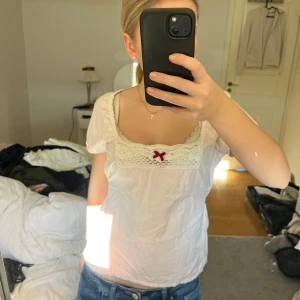Ljus rosa blus med spets och rosett - Charmig vit blus med spetsdetaljer och en söt röd rosett framtill. Blusen har korta ärmar och ett brunt knytband i midjan för en fin passform. Kommer från odd molly och har knappt använts! 