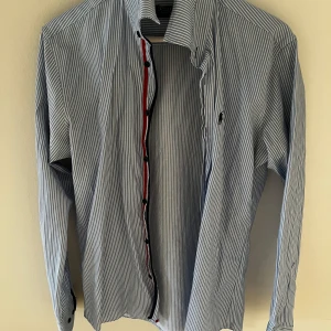 Randig skjorta med knappar Ralph Lauren  - Ralph Lauren Snygg randig skjorta i blått och vitt med knappar framtill. Perfekt för en stilren look. Skjortan har långa ärmar och en klassisk krage.