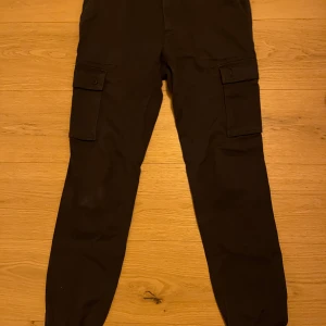 Svarta cargobyxor från Pier One - Snygga svarta cargobyxor från Pier One med praktiska fickor på sidorna. Byxorna har en normal passform och är perfekta för en avslappnad stil. Tillverkade i ett slitstarkt material med knappar på fickorna för extra säkerhet.