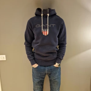 Mörkblå hoodie från GANT - Säljer en snygg mörkblå hoodie från GANT med ett stilrent tryck och logga på bröstet. Tröjan har en praktisk känguruficka och justerbar huva med vita snören. Vid funderingar är det bara att skriva// merch Åled