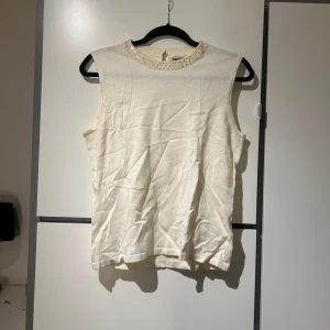 Vit ärmlös topp med pärldetaljer - Säljer en stilren vit ärmlös topp med  pärldetaljer runt halsringningen. Toppen är i ett mjukt material och har en knapp i nacken för enkel på- och avtagning.