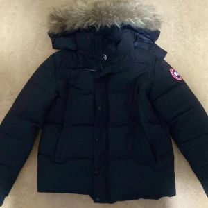 Svart dunjacka från Canada Goose  /priset går o diskutera - Säljer en svart dunjacka från Canada Goose med en avtagbar huva och pälsdetaljer. Jackan har en broderad logotyp på ärmen och stängs med dragkedja och knappar. Perfekt för kalla vinterdagar.