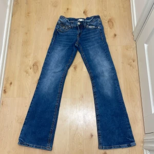 Blå bootcut jeans - Snygga blå bootcut jeans med klassisk femficksdesign och knappgylf. Perfekta för en avslappnad stil med en touch av retro. De har en lätt tvättad look som ger en cool vintagekänsla.