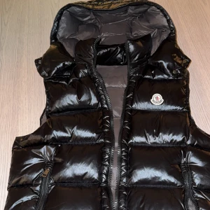 Svart dunväst från Moncler  - Snygg svart dunväst från Moncler med glansig finish och dragkedja. Västen har en praktisk huva och Moncler-logga på bröstet. Perfekt för kyligare dagar när du vill hålla stilen. Tag ingår. Storlek S/M.