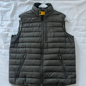 Parajumpers bäst - Säljer en snygg grå dunväst från Parajumpers. Västen har en dragkedja framtill och en hög krage med en gul detalj. Perfekt för kyligare dagar och ger en stilren look. Den är i mycket bra skick förutom några mindre fläckar som knappt syns se bild 2
