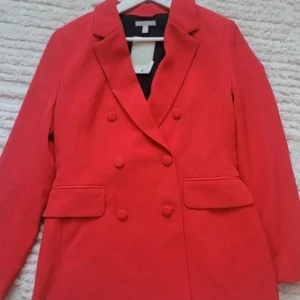Röd kavaj från H&M - Snygg röd kavaj från H&M med dubbelknäppning och klassisk krage. Perfekt för att ge en färgklick till din outfit. Tillverkad i polyester med två fickor framtill. En stilren och tidlös design som funkar till många tillfällen.