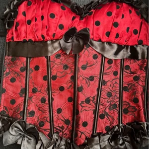 Fin korsett i Moulin Rouge-stil! - Jättefin korsett i röd satin med svart spets med dragkedja i sidan, justerbara axelband, rygglapp, mjuka skenor fram och snörning bak som kan justeras flera dm. Mått: H: 40 cm (exkl. axelband) 80 cm runtom på innersta snörning med utrymme för extra 20 cm. Perfekt skick.
