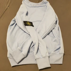 Ljusblå tröja från Stone Island - Säljer en stilren ljusblå tröja från Stone Island. Tröjan har långa ärmar och ribbade muddar vid ärmslut och nederkant. Perfekt för en avslappnad stil. 🌀