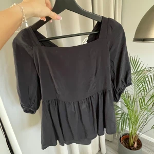 Svart blus från Aware - Snygg svart blus från Aware med fyrkantig halsringning och puffiga trekvartsärmar. Blusen har en knytning i ryggen och en volangdetalj nedtill som ger en fin siluett. Perfekt för en stilren look.