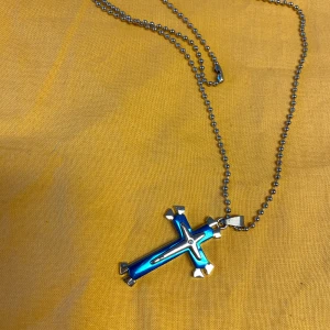 Halsband med korshänge i olika färger - Säljer flera halsband med korshängen. Ett har ett blått kors med silverdetaljer på en kulkedja. Ett annat har ett svart kors med silverdetaljer. Det tredje har ett dekorativt kors med en röd sten i mitten på en svart rem. Perfekt för att ge din stil en unik touch.  Helt ny, 125kr/st 