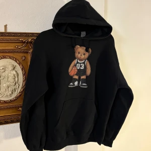 Fashion bear hoodie - Säljer en svart hoodie med ett gulligt nallemotiv som håller en basketboll och har nummer 03 på tröjan. Hoodien har en klassisk passform med långa ärmar och en praktisk ficka framtill. Perfekt för en avslappnad stil! Storlek M.