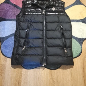 Svart dunväst från Moncler - Snygg svart dunväst från Moncler med glansig finish. Västen har en dragkedja framtill och två praktiska fickor med dragkedjor. Perfekt för kyligare dagar när du vill hålla stilen. Men lite mer åt M hållet