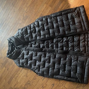 Svart dunväst från Everest - Säljer en svart dunväst från Everest. Västen har en quiltad design och dragkedja framtill. Perfekt för kyligare dagar när du vill hålla värmen utan att bära en jacka. Märket Everest är känt för sina funktionella och stilrena plagg.