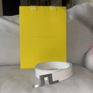 Vitt bälte från J.Lindeberg - Stilrent vitt bälte från J.Lindeberg med en unik silverfärgad spänne. Perfekt för att ge en modern touch till din outfit. Bältet är tillverkat i ett elegant material och har en diskret logotyp på insidan.