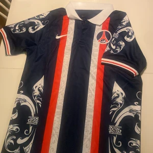 Retro PSG tröja - Snygg mörkblå fotbollströja från Nike med vita och röda ränder samt dekorativa mönster på ärmarna. Tröjan har en vit krage och korta ärmar med röd-vit rand vid ärmslutet. Perfekt för fotbollsfans som vill sticka ut på planen!