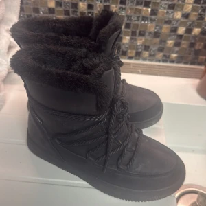 Svarta moon boots-like med pälsfoder - Säljer ett par svarta boots  moon-boots-stil med mjukt pälsfoder och snörning. Perfekta för kalla vinterdagar med sin varma och bekväma design.