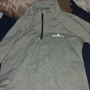 Grå tröja med dragkedja från Ronhill - Säljer en grå tröja från Ronhill med halv dragkedja och hög krage. Perfekt för löpning eller träning. Tröjan har en sportig design och är tillverkad i ett bekvämt material.