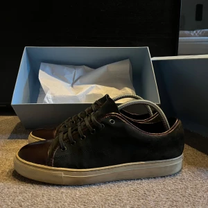 Svarta sneakers med bruna detaljer - Snygga svarta sneakers med bruna detaljer och snörning. Skorna har en vit sula och är perfekta för en avslappnad stil. De kommer i originalkartong. Röd CapToe