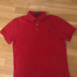 Röd pikétröja från Ralph Lauren - Snygg röd pikétröja från Ralph Lauren i slim fit. Tröjan har en klassisk krage med knappar och det ikoniska broderade logotypen på bröstet. Perfekt för en stilren look.