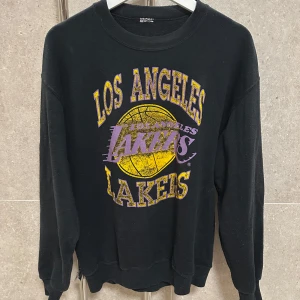 Svart Los Angeles Lakers sweatshirt - Cool svart sweatshirt med Los Angeles Lakers tryck i gult och lila. Tröjan har långa ärmar och en rund halsringning. Perfekt för basketfans som vill visa sitt stöd för laget.