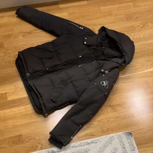 Svart dunjacka från Moose Knuckles -  svart dunjacka från Moose Knuckles. Jackan har en avtagbar huva och flera fickor med dragkedjor. Perfekt för kalla dagar med sin quiltade design och långa ärmar. Vit päls finns till jackan.