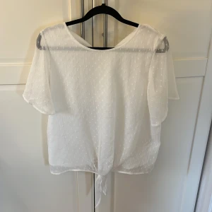 Vit blus med knyt - Säljer en söt vit blus med korta ärmar och knytning i nederkant. Blusen har ett diskret prickigt mönster och är tillverkad i ett lätt och luftigt material. Perfekt för varmare dagar!