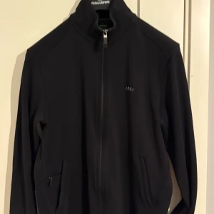 Svart sweatshirt från Boss - Snygg svart jacka från Boss med dragkedja framtill och diskret logga på bröstet. Jackan har långa ärmar och praktiska fickor med dragkedja. Perfekt för en stilren look.