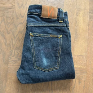 Mörkblåa Nudie Jeans - Säljer ett par mörkblåa Nudie Jeans i modellen "Grim Tim", storlek W32 L34. Tillverkade i 100% bomull med en rak passform, vilket ger en stilren och bekväm look. Jeansen är i ett mycket bra skick och är i väldig bra kvalitiet. Nypris 1600 kr.