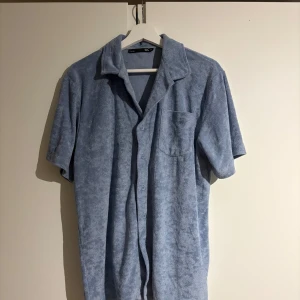 Blå frottéskjorta - Säljer en blå frottéskjorta med korta ärmar och knappar framtill. Skjortan har en avslappnad passform och en bröstficka. Perfekt för en ledig stil.
