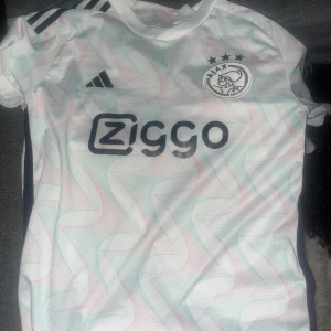 Fotbollströja  - Säljer en snygg vit och rosa fotbollströja från Adidas med Ajax-logga och Ziggo sponsortryck. Tröjan har korta ärmar och ett unikt mönster i ljusa färger. Perfekt för fotbollsfans som vill sticka ut på planen!