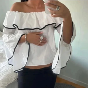 Svart offshoulder blus med volangdetaljer - Trendig svart offshoulder blus med vita volangdetaljer. Perfekt för en stilren look. Blusen har vida ärmar och en luftig passform.🤍🤍första bilden är lånad men liknar fast i vit