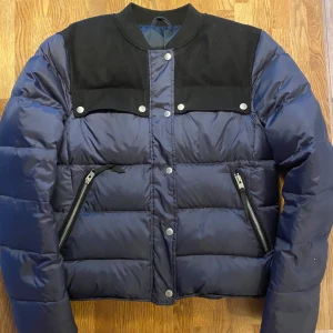 Blå dunjacka från J.Lindeberg - Säljer en snygg blå dunjacka från J.Lindeberg med svarta detaljer på axlarna. Jackan har dragkedjor på fickorna och knappar framtill.