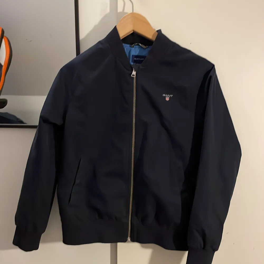 Snygg mörkblå bomberjacka från GANT med dragkedja framtill och ribbade muddar. Jackan har en stilren design med GANT-logga på bröstet och ett ljust foder med logotyp inuti. Perfekt för en casual look. Skick 10/10. Inga hål eller skrapor.. Takit.