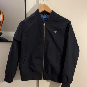 Mörkblå bomberjacka från GANT - Snygg mörkblå bomberjacka från GANT med dragkedja framtill och ribbade muddar. Jackan har en stilren design med GANT-logga på bröstet och ett ljust foder med logotyp inuti. Perfekt för en casual look. Skick 10/10. Inga hål eller skrapor.