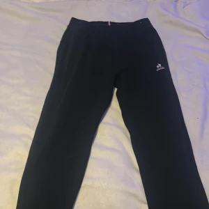 Svarta mjukisbyxor från Le Coq Sportif - Säljer ett par svarta mjukisbyxor från Le Coq Sportif med deras ikoniska logga broderad på benet. Byxorna har en bekväm passform och elastisk midja med en trikolor-detalj. Perfekta för en avslappnad stil.