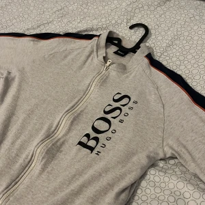 Grå hoodie från Hugo Boss - Säljer en stilren grå hoodie från Hugo Boss med dragkedja och tryck på bröstet. Den har långa ärmar med en mörkblå och orange rand längs ärmarna. Perfekt för en avslappnad look.