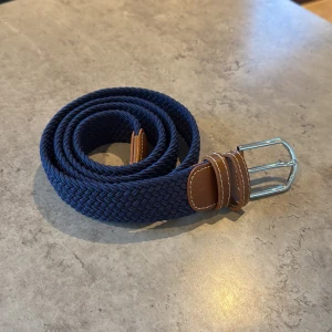 Blått flätat bälte med bruna detaljer - Snyggt blått flätat bälte med bruna skinnliknande detaljer och en silverfärgad spänne. Perfekt för att ge en stilren touch till din outfit. Bältet har en klassisk design som passar till många olika stilar. Bältet är även 120cm