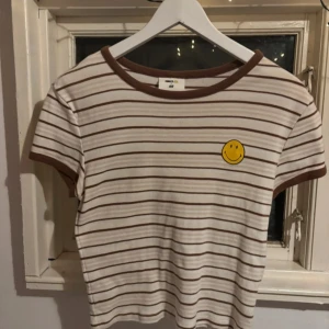 Randig kortärmad topp med smiley - Säljer en söt randig topp från H&M i beige och brunt med en glad smiley på bröstet. Toppen har korta ärmar och en rund halsringning med bruna kanter. Mycket sparsamt använd, inga defekter.