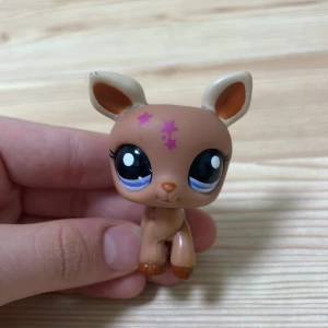 Littlest Pet Shop figur - Söt Littlest Pet Shop figur i form av ett rådjur med stora blå ögon och rosa stjärnor på huvudet. Figuren är brun med ljusare detaljer på öronen och svansen. Perfekt för samlare eller som leksak.
