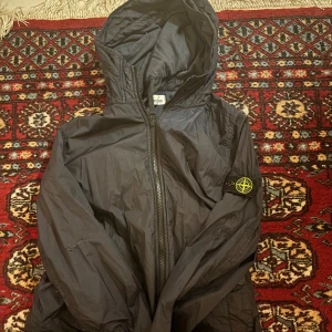 Svart vindjacka från Stone Island Junior - Säljer en svart vindjacka från Stone Island Junior. Jackan har en huva och dragkedja framtill. Den är tillverkad i ett lätt syntetmaterial och har en ikonisk logga på ärmen.