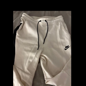 Vita mjukisbyxor från Nike - Snygga vita mjukisbyxor från Nike med svart snörning i midjan. Byxorna har en avslappnad passform och är perfekta för en bekväm stil. Nike-loggan är broderad på sidan.