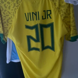 Vinicius fotbollströja  - Snygg gul Brasilien fotbollströja från Nike med VINI JR och nummer 20 på ryggen. Tröjan har ett leopardmönster och korta ärmar med gröna detaljer vid ärmslut och krage. Perfekt för fotbollsfans som vill visa sitt stöd för laget! I storlek M
