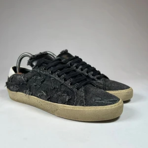 Svarta sneakers från Saint Laurent - Snygga svarta sneakers från Saint Laurent med en unik, sliten look. Skorna har en beige sula och är tillverkade i ett denimliknande material med fransiga detaljer. De har klassisk snörning och en vit hälflik som kontrasterar mot det svarta.