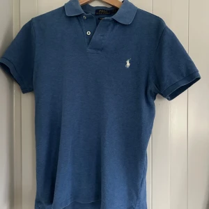 Blå pikétröja från Ralph Lauren - Säljer en stilren blå pikétröja från Ralph Lauren i custom slim fit. Tröjan har korta ärmar och en klassisk krage med knappar. Den ikoniska loggan är broderad på bröstet. Perfekt för en avslappnad men ändå snygg look.