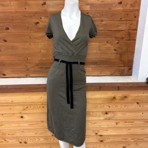 Olivgrön omlottklänning med bälte - Snygg olivgrön omlottklänning med korta ärmar och ett svart knytbälte i midjan. Klänningen har en v-ringad design och är perfekt för en stilren look. Den är gjord i ett mjukt material som ger en bekväm passform.