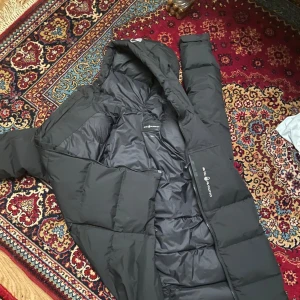 Svart dunjacka från sail racing - Säljer en svart dunjacka från sail racing. Jackan har en dragkedja framtill och en huva för extra värme. Perfekt för kalla dagar med sin quiltade design. Den är långärmad och har en stilren look. Har ett litet märke där bak men märks knappast 